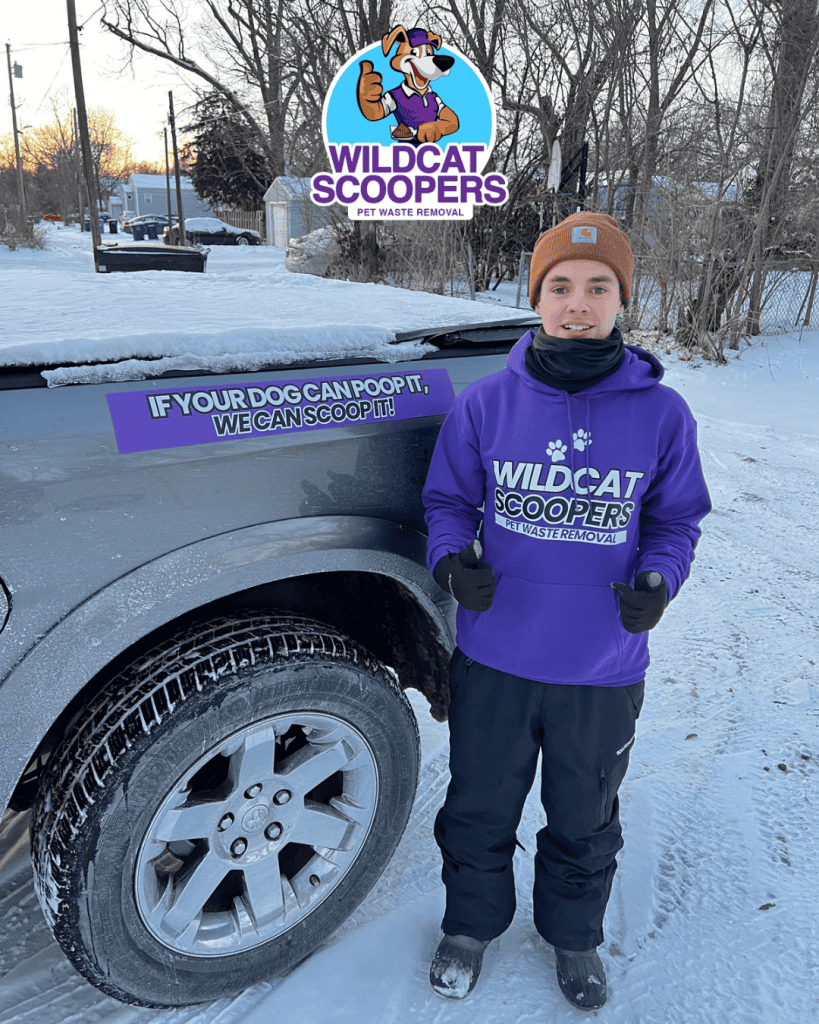 A person dressed in winter attire is standing next to a dark-colored vehicle with a Wildcat Scoopers logo on the door. They are wearing a purple hoodie with the same logo and giving a thumbs-up. Snow covers the ground, and houses are visible in the background. A person dressed in winter attire is standing next to a dark-colored vehicle with a Wildcat Scoopers logo on the door. They are wearing a purple hoodie with the same logo and giving a thumbs-up. Snow covers the ground, and houses are visible in the background.