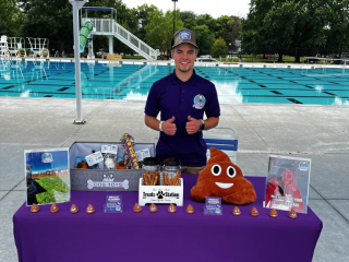 A person stands behind a purple table covered with informational displays, plush toys, and small items. They are giving two thumbs up. The setting appears to be an outdoor pool area with trees in the background. A person stands behind a purple table covered with informational displays, plush toys, and small items. They are giving two thumbs up. The setting appears to be an outdoor pool area with trees in the background.