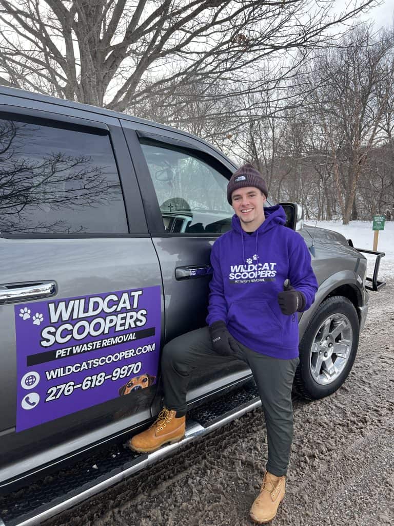 A person wearing a purple hoodie and a black beanie stands on the step of a black truck with “Wildcat Scoopers: Pet Waste Removal” signage. They are smiling and giving a thumbs-up, with a snowy landscape in the background. A person wearing a purple hoodie and a black beanie stands on the step of a black truck with "Wildcat Scoopers: Pet Waste Removal" signage. They are smiling and giving a thumbs-up, with a snowy landscape in the background.