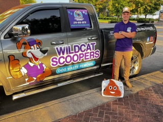 A person in a purple shirt and cap stands beside a truck with “Wildcat Scoopers” branding. They’re holding a large bag with a poop emoji. The truck is parked on a street with buildings and trees in the background. A person in a purple shirt and cap stands beside a truck with "Wildcat Scoopers" branding. They're holding a large bag with a poop emoji. The truck is parked on a street with buildings and trees in the background.