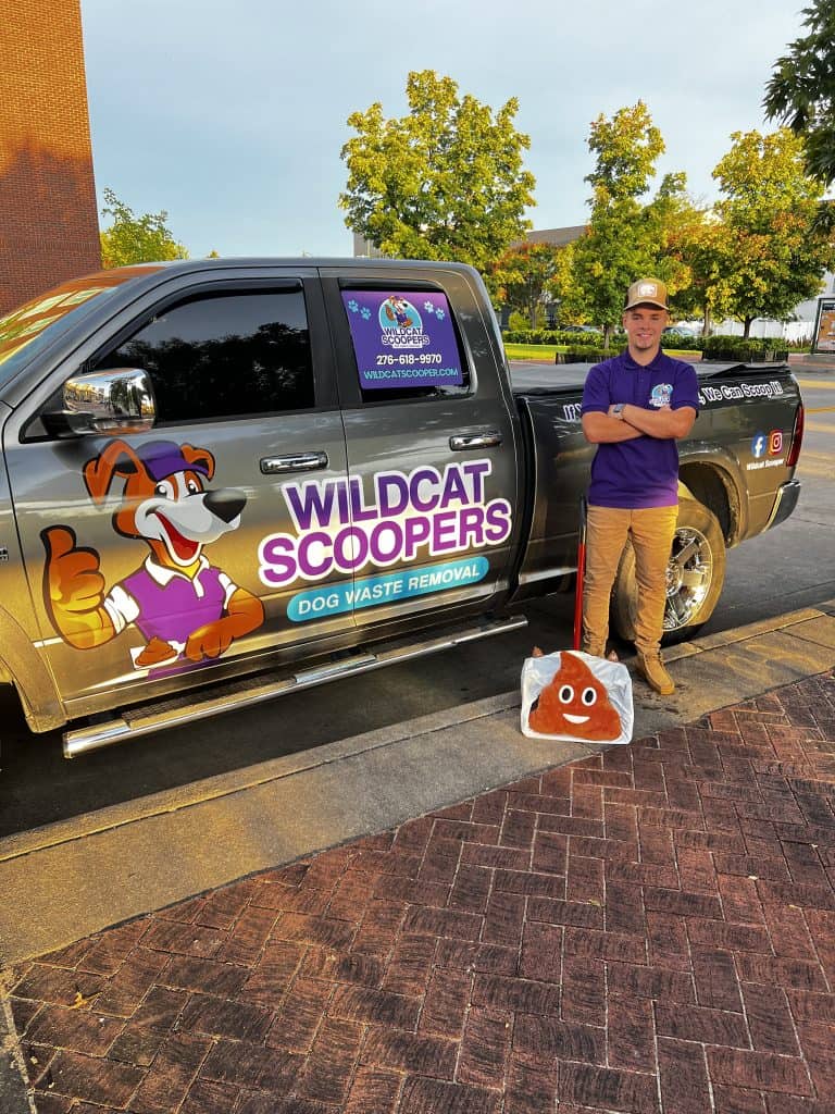 A person in a purple shirt and cap stands beside a truck with “Wildcat Scoopers” branding. They’re holding a large bag with a poop emoji. The truck is parked on a street with buildings and trees in the background. A person in a purple shirt and cap stands beside a truck with "Wildcat Scoopers" branding. They're holding a large bag with a poop emoji. The truck is parked on a street with buildings and trees in the background.
