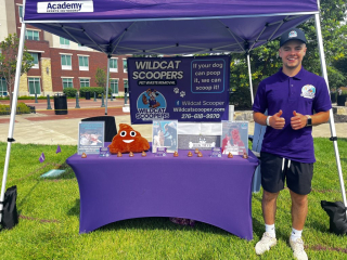 A person stands under a tent with a Wildcat Scoopers banner. The table has a plush emoji poop, brochures, and small figurines. The person is wearing a purple shirt and cap, giving two thumbs up. A sign reads “If your dog can poop it, we can scoop it!” in front of a grassy area. A person stands under a tent with a Wildcat Scoopers banner. The table has a plush emoji poop, brochures, and small figurines. The person is wearing a purple shirt and cap, giving two thumbs up. A sign reads "If your dog can poop it, we can scoop it!" in front of a grassy area.
