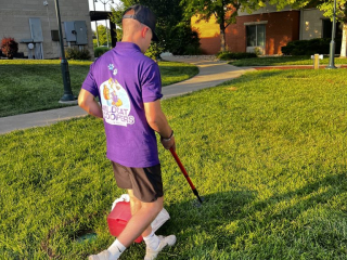 A man in a purple “Wildcat Hoopers” shirt and black shorts uses a picker tool to collect trash on a grassy area in the bright outdoor sunshine. He carries a red container for the collected items. Trees and buildings are in the background. A man in a purple "Wildcat Hoopers" shirt and black shorts uses a picker tool to collect trash on a grassy area in the bright outdoor sunshine. He carries a red container for the collected items. Trees and buildings are in the background.