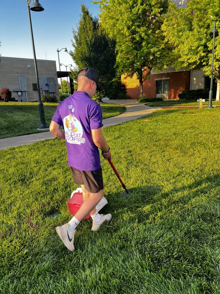 A man in a purple “Wildcat Hoopers” shirt and black shorts uses a picker tool to collect trash on a grassy area in the bright outdoor sunshine. He carries a red container for the collected items. Trees and buildings are in the background. A man in a purple "Wildcat Hoopers" shirt and black shorts uses a picker tool to collect trash on a grassy area in the bright outdoor sunshine. He carries a red container for the collected items. Trees and buildings are in the background.