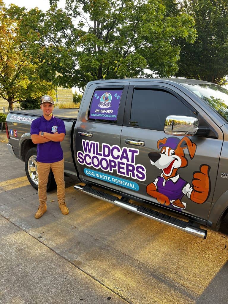 A person wearing a purple shirt stands next to a truck with "Wildcat Scoopers Dog Waste Removal" and a cartoon dog logo. The truck is gray, parked outdoors in a sunny area with trees in the background.
