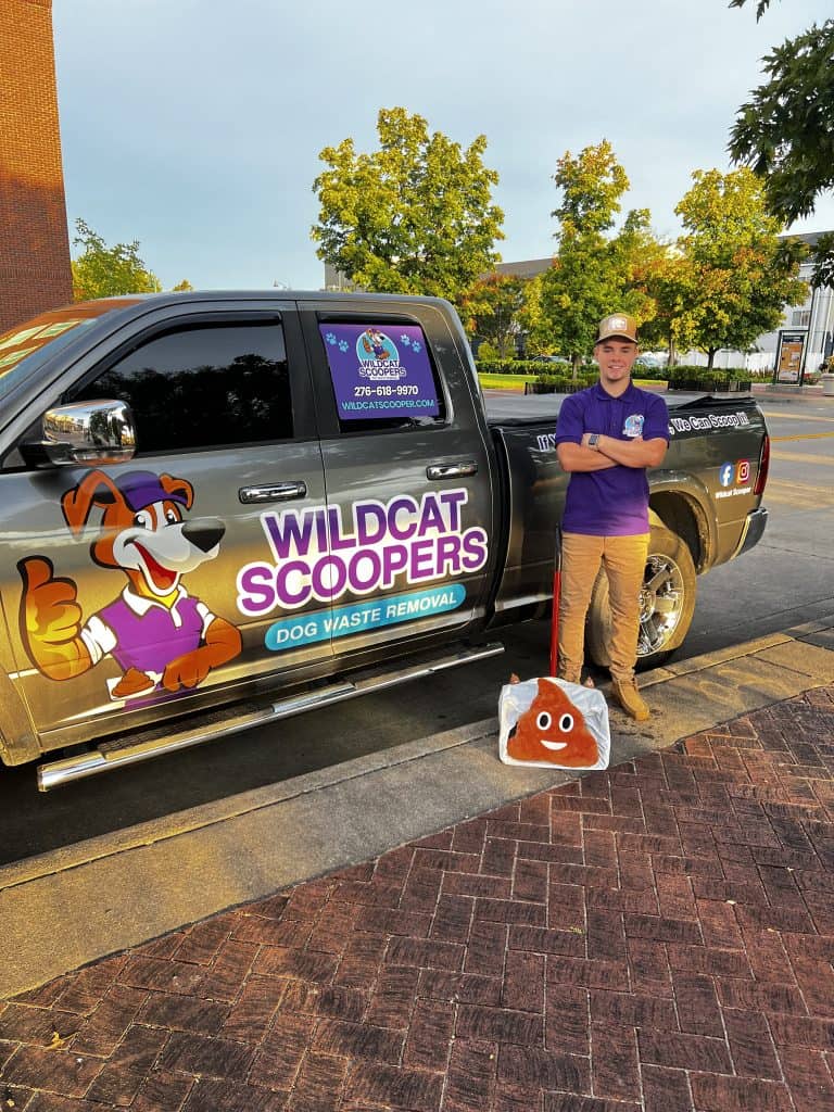 A person wearing a purple shirt and cap stands next to a truck with "Wildcat Scoopers Dog Waste Removal" on it. They hold a large novelty poop emoji scoop. The background shows trees and brick pavement.
