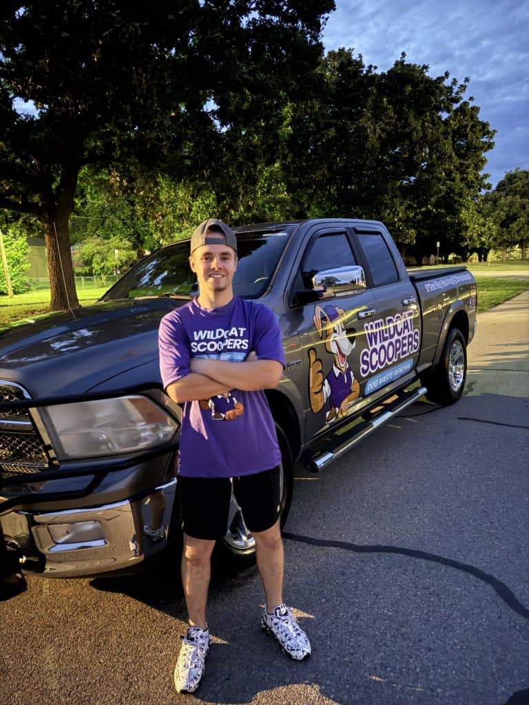 A young man in a purple "Wildcat Scoopers" t-shirt and cap stands smiling in front of a black pickup truck with the "Wildcat Scoopers" logo and mascot on the door, parked on a sunny street by green trees.