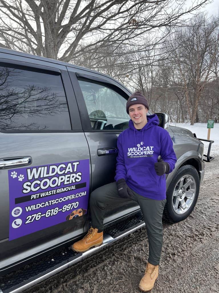 A smiling person in a purple “Wildcat Scoopers” hoodie stands next to a truck with a “Wildcat Scoopers Pet Waste Removal” sign, parked on a snowy road with bare trees in the background.