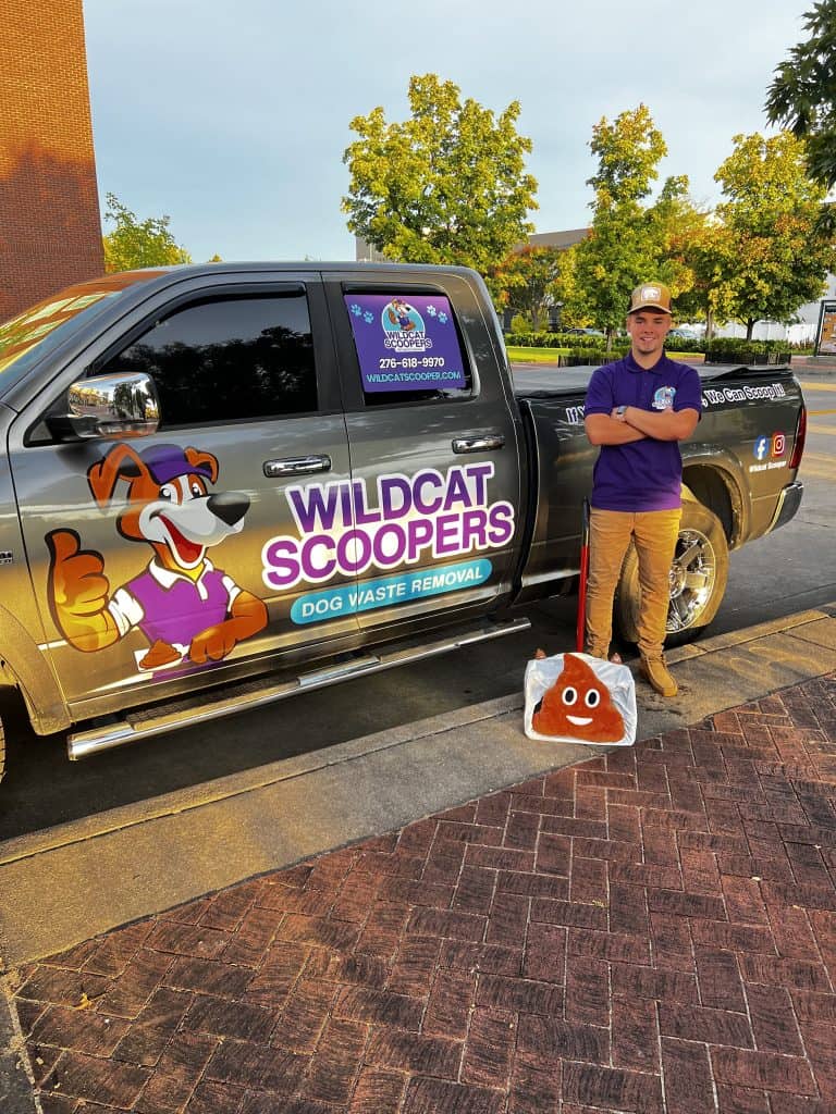 A man in a purple polo and cap stands next to a truck labeled "Wildcat Scoopers Dog Waste Removal," holding a large poop emoji bag. The truck features a cartoon dog mascot and colorful signage. Trees and buildings are in the background.