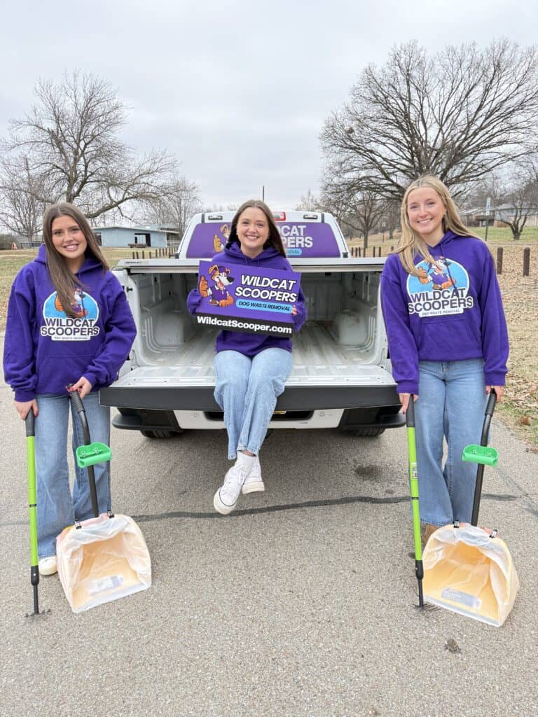 Three smiling women in "Wildcat Scoopers" sweatshirts pose by a pickup truck. Two hold litter scoopers, and one sits on the truck bed holding a company sign. Leafless trees and cloudy skies are in the background.