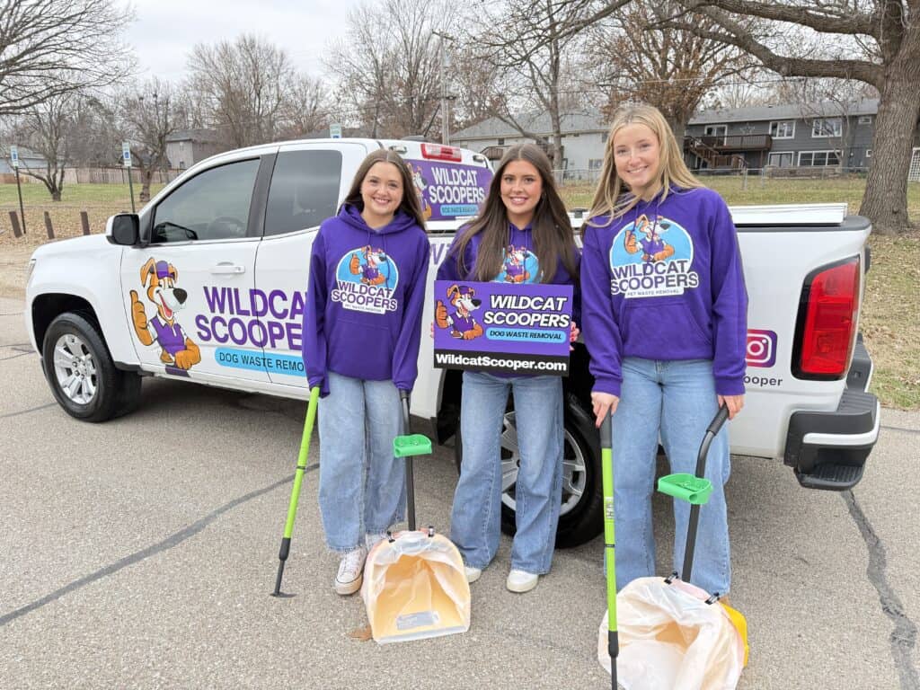 Three smiling young women in purple "Wildcat Scoopers" hoodies stand in front of a branded pickup truck, holding poop scoopers and waste bags. One holds a company sign. The background shows trees and houses.